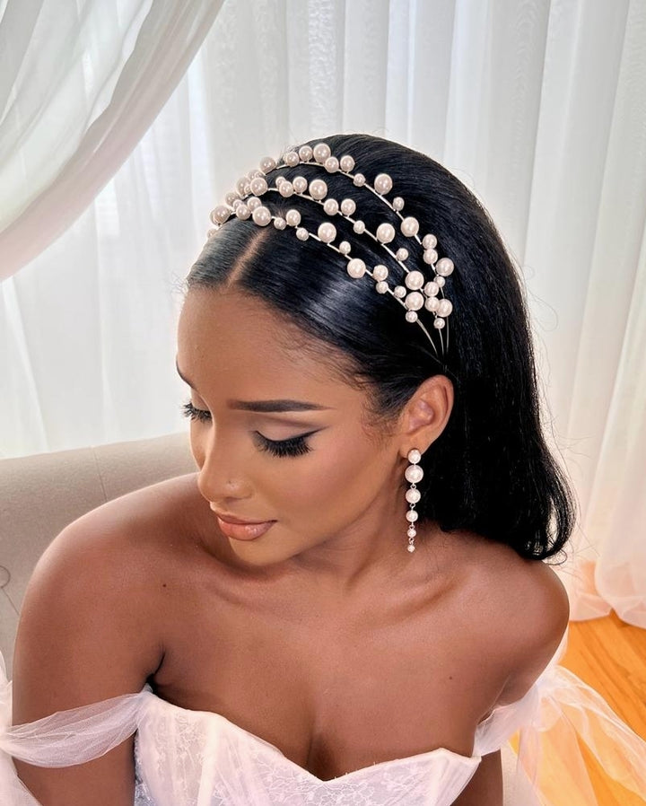 A woman with smooth, dark hair wears Pearl Drop Earrings style no. E214 and a pearl-studded headband, paired with elegant makeup and an off-the-shoulder white dress as she sits by sheer curtains in soft natural light.