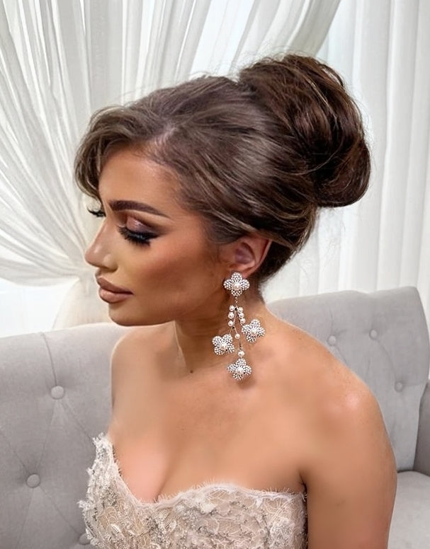 A woman with an elegant updo and dramatic makeup wears the Pearl Blossom Chandelier Earrings (style no. E217) and a strapless textured gown, sitting on a light grey tufted sofa in front of sheer white curtains.