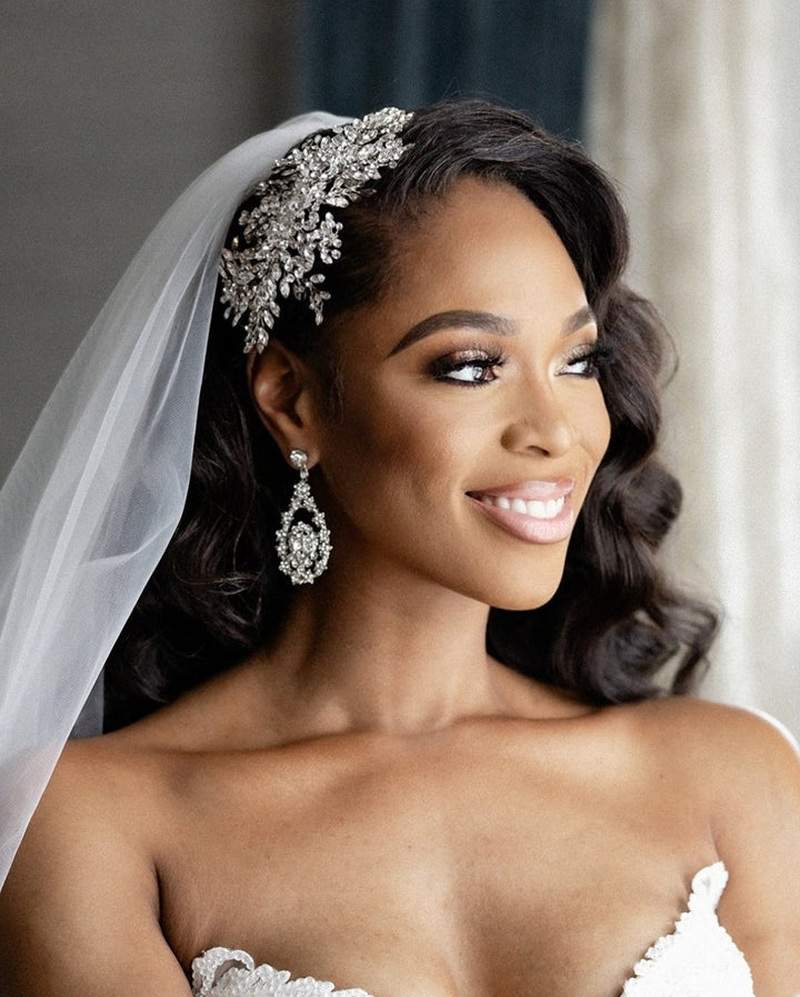 Bride in voluminous curls and veil wearing Swarovski crystal chandelier earrings, paired with a dramatic crystal bridal comb for a glamorous wedding look.

