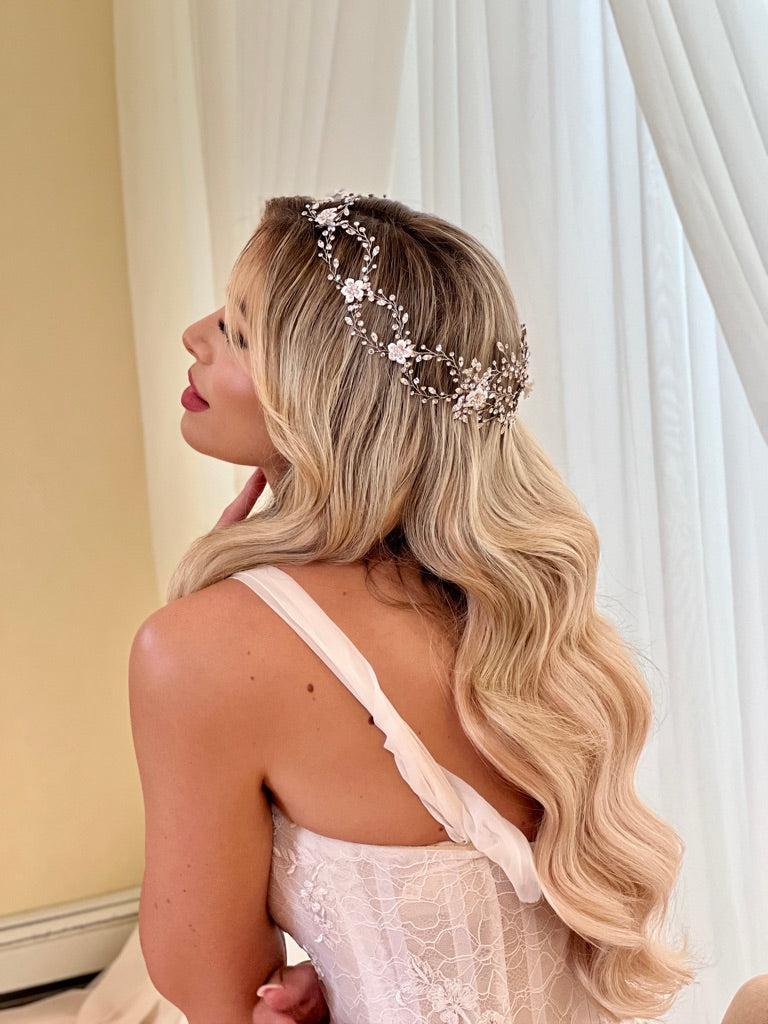 A woman with long, wavy blonde hair sits gracefully in front of white curtains, adorned with the Enchanting Swarovski Crystal and Flower Wedding Halo (style no. 242713). Wearing a white, lacy dress, she tilts her head slightly back and gently touches her chin with her hand.