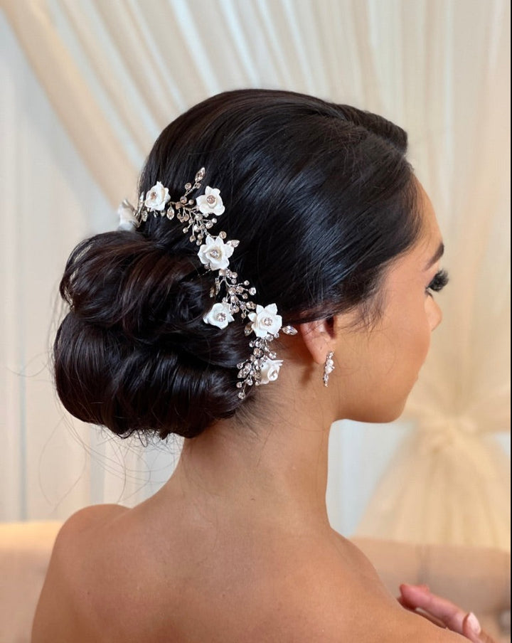 A woman with dark hair in an elegant bun, adorned with the Romantic Flower and Swarovski Crystal Hair Vine style no. 242720 featuring white ceramic flowers and crystal accents, wears delicate drop earrings against a backdrop of soft cream drapes.