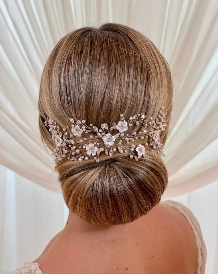 A woman with a low, elegant chignon hairstyle is adorned with the Delicate Pearl and Porcelain Flower Vine (style no. 242712). The Swarovski crystal bridal hair vine features white flowers and pearls against a soft, white curtain background.