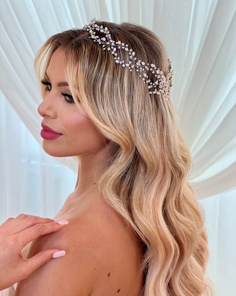 A woman with long, wavy blonde hair wears the Romantic Crystal and Pearl Wedding Halo style no. 242675. She has pink lipstick, a light skin tone, and a neutral manicure as she stands gracefully in front of a sheer white curtain.
