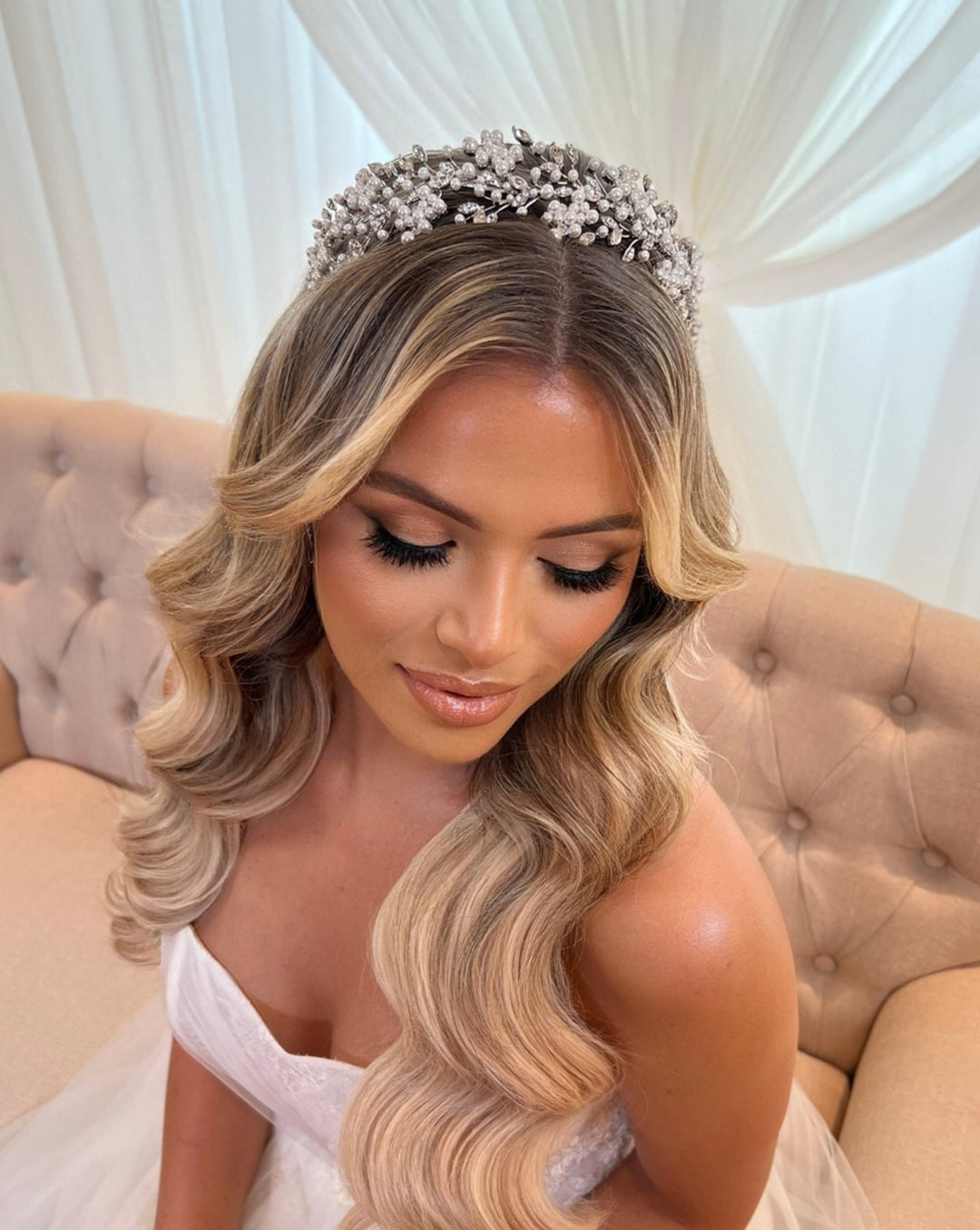 A bride with long, wavy blonde hair and elegant makeup sits on a beige tufted couch, wearing a white strapless gown and the Shimmering Cluster Headband style no. 242685. Draped white fabric enhances the backgrounds ethereal charm.