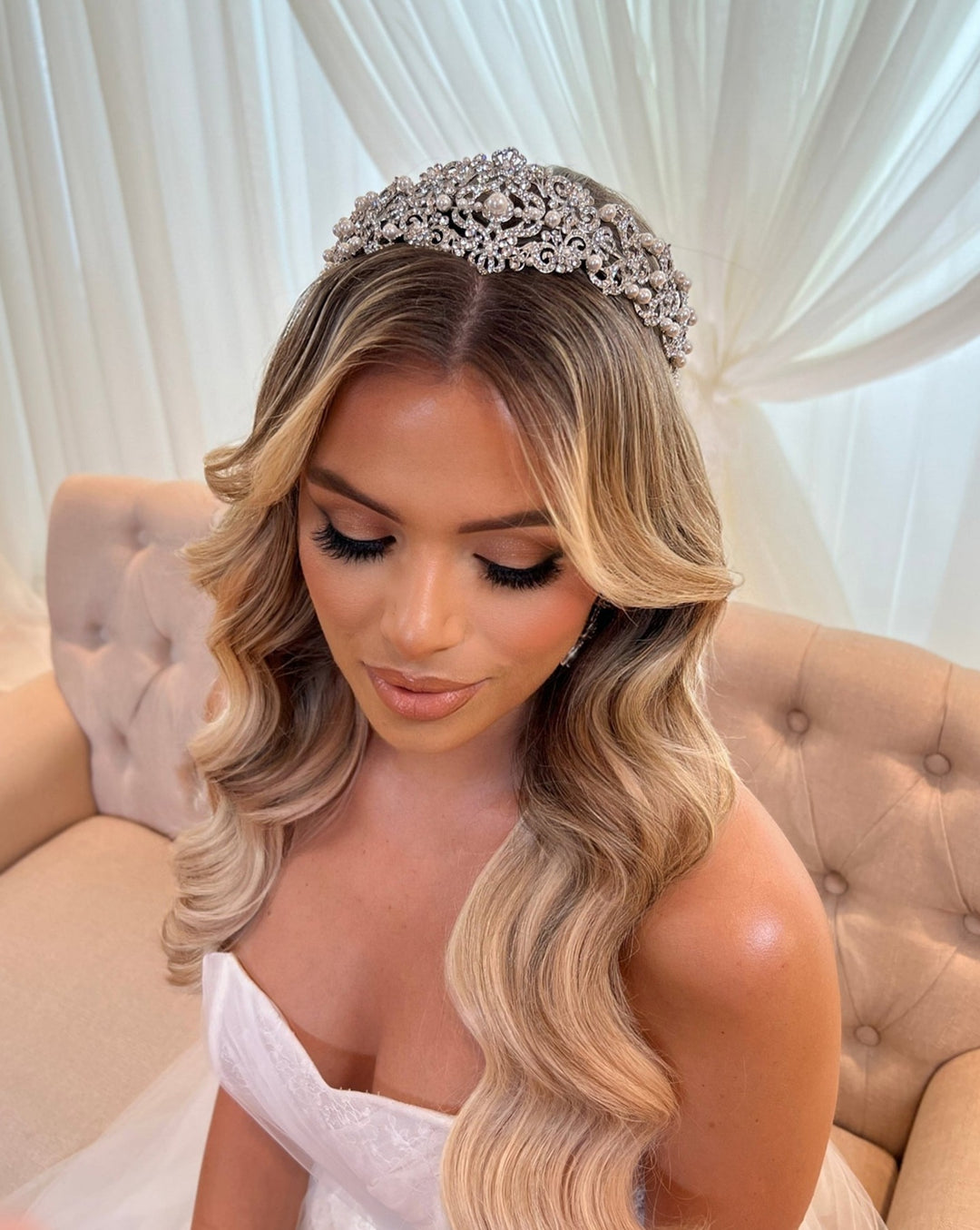 A woman with long, wavy blonde hair sits on a beige couch, wearing a Vintage Inspired Swarovski Crystal Headband style no. 80107 and an off-shoulder white dress. Her eyes are closed, and a sheer curtain is draped behind her.