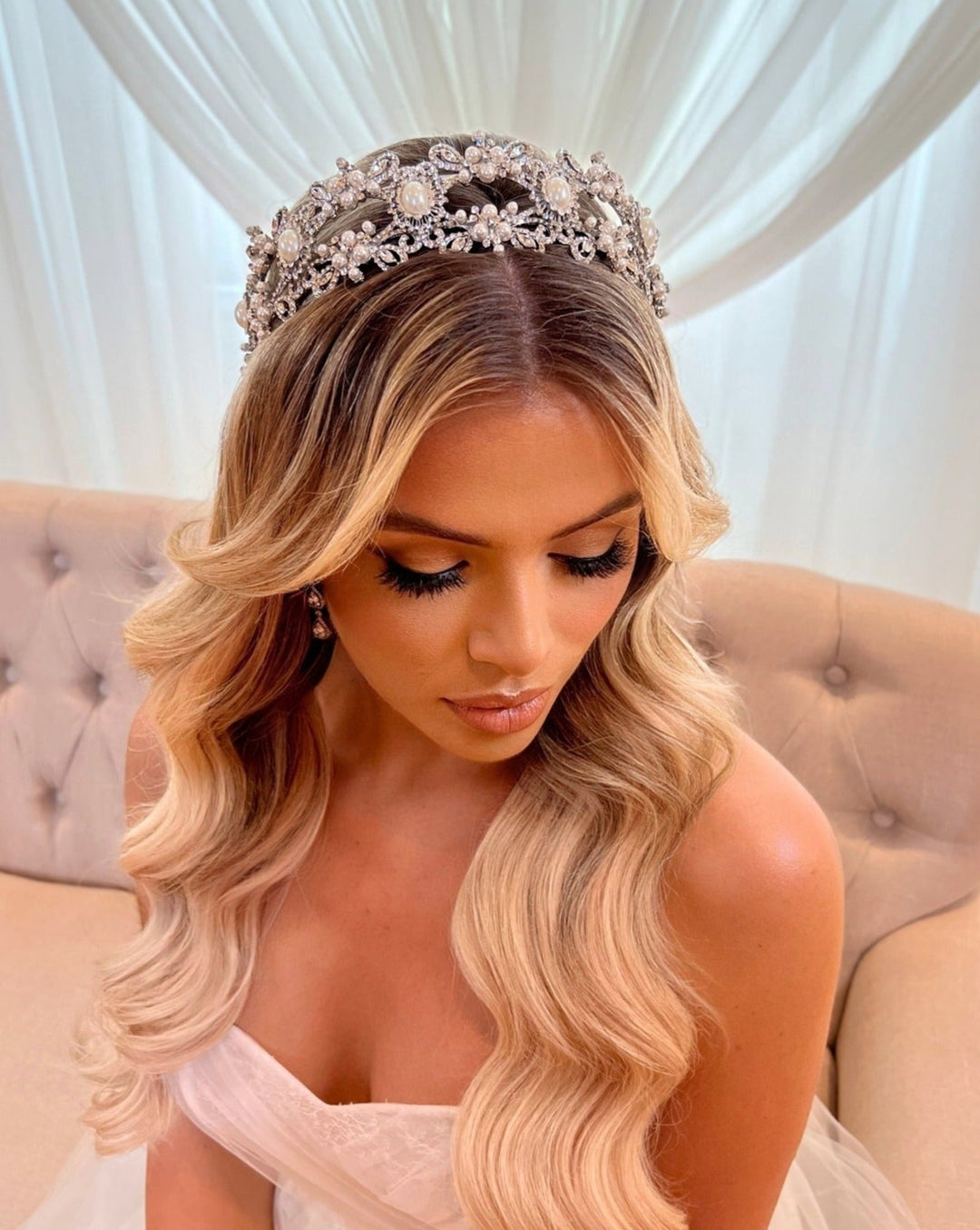 A woman with long, wavy blonde hair wears a sparkling Vintage Inspired Pearl and Swarovski Crystal Headband style no. 80055 as she sits on a light beige tufted sofa. Her dramatic eye makeup stands out against the backdrop of soft, white drapery as she looks downward.
