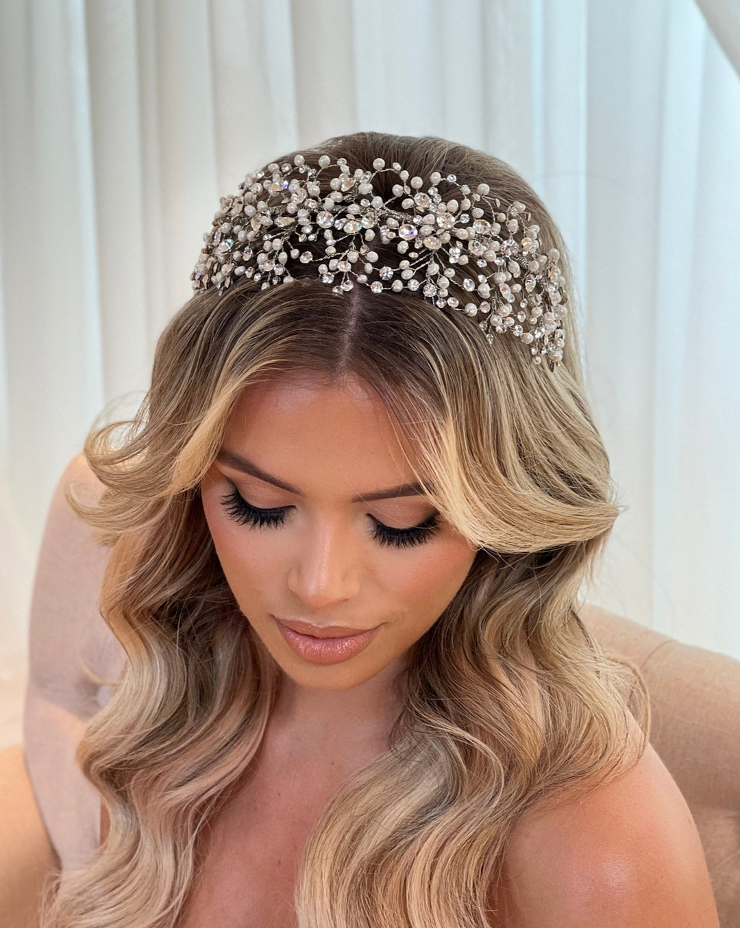 A person with long, wavy hair sits, eyes closed, wearing the Classy Bridal Headband style no. 242676. Their subtle makeup complements their elegant look amidst a softly blurred background, enhancing the regal aura of the ornate headpiece.