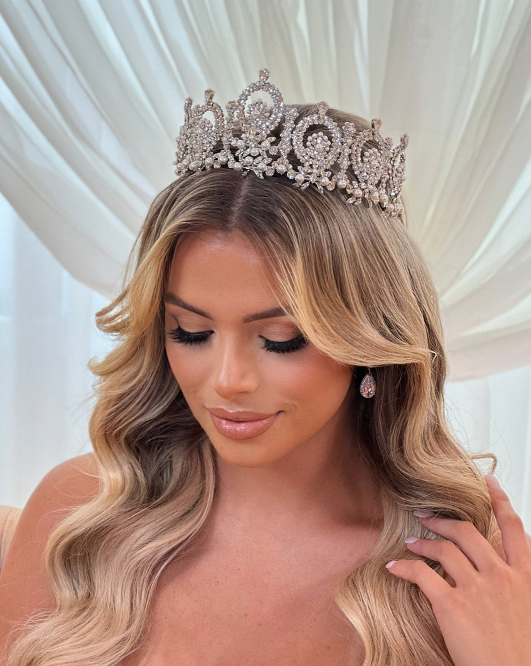 A woman with long, wavy blonde hair wears a Regal Wedding Crown, style no. 80015. Her eyes are closed, highlighting her makeup and drop earrings. She gently touches the Swarovski bridal crown against a soft, creamy backdrop.