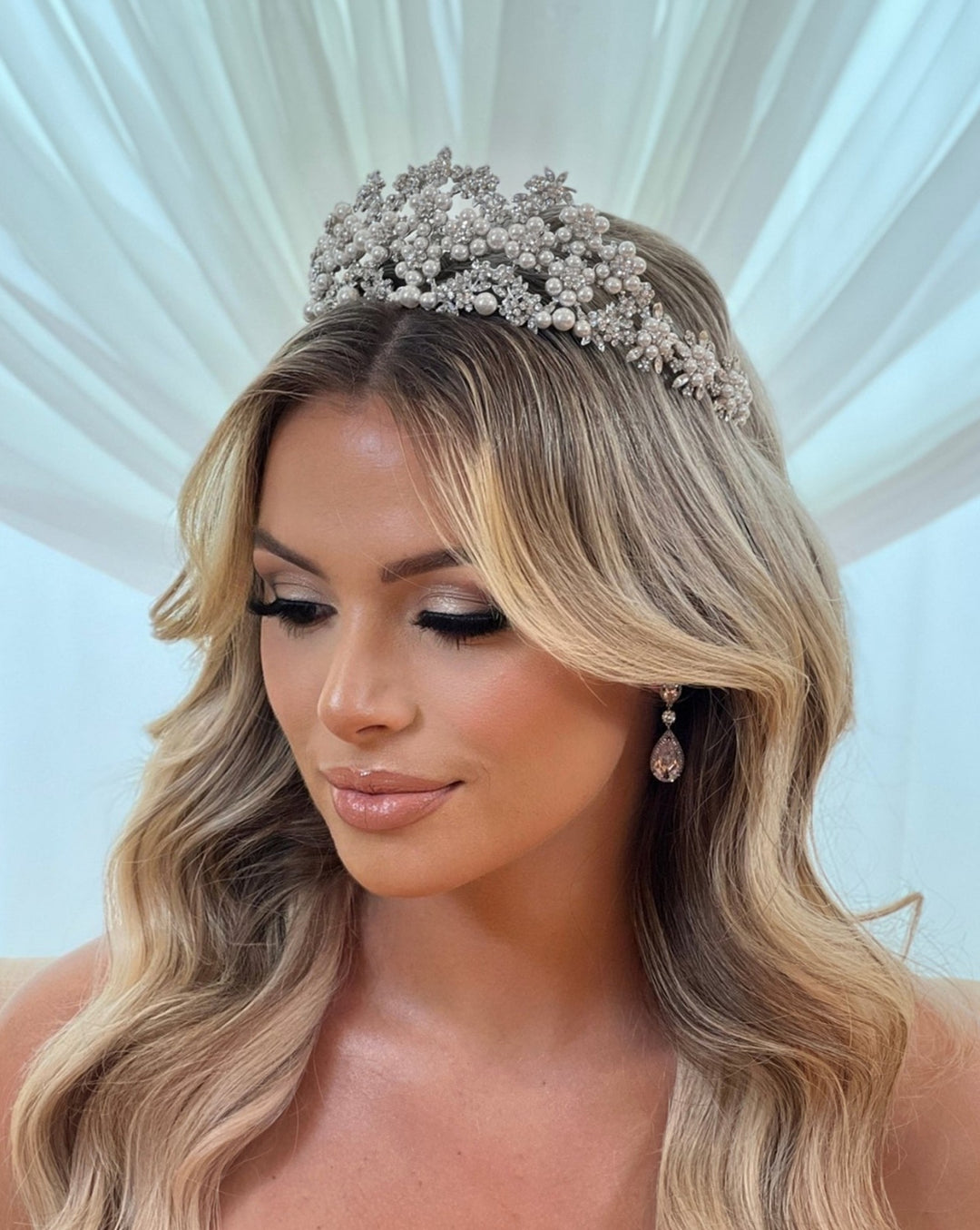 A woman with wavy blonde hair wears the Pearl and Swarovski Crystal Bridal Tiara (style no. 80044) and drop earrings. Her eyes are closed as she stands before a soft, flowing white backdrop, exuding an elegant bridal appearance.