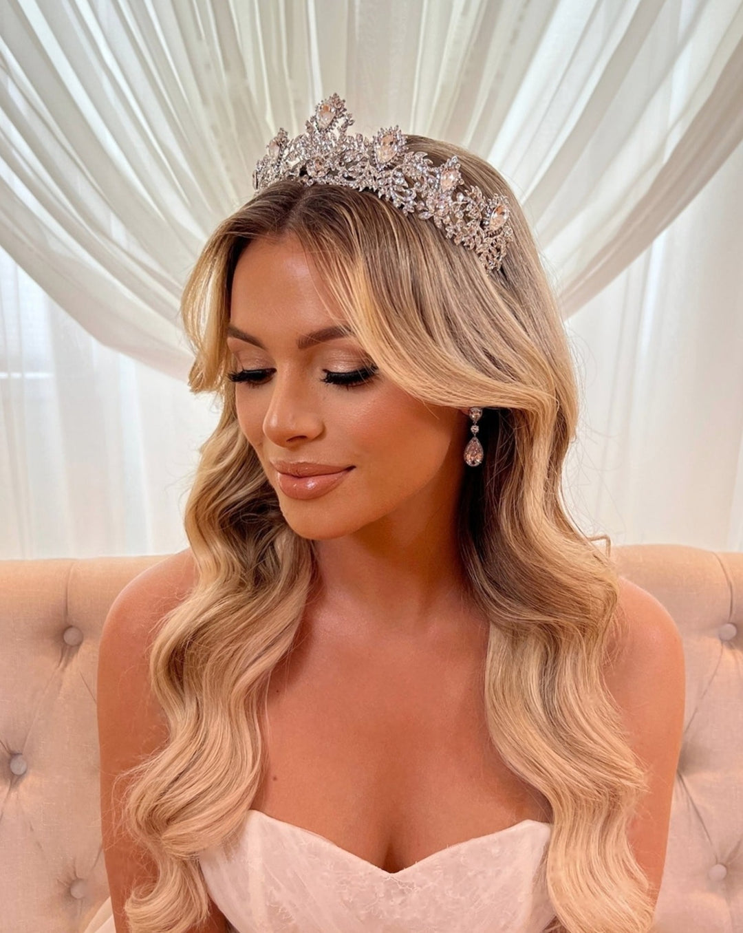 A woman in a strapless wedding dress sits on a beige couch. Her long, wavy blonde hair is elegantly adorned with the Glamorous Swarovski Crystal Tiara style no. 80100, and she wears drop bridal earrings, gazing downward against sheer white curtains.