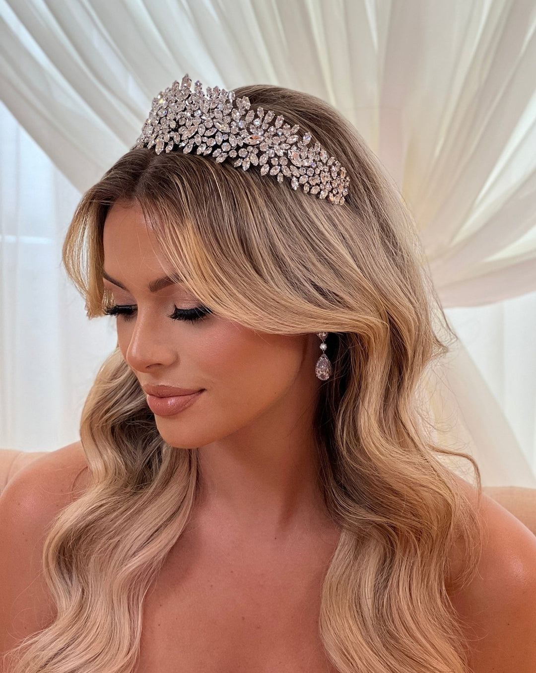 A woman with long wavy blonde hair wears a Dazzling Swarovski Crystal Bridal Headband (style no. 7900806) and teardrop earrings. Her eyes are closed against a backdrop of sheer white curtains.