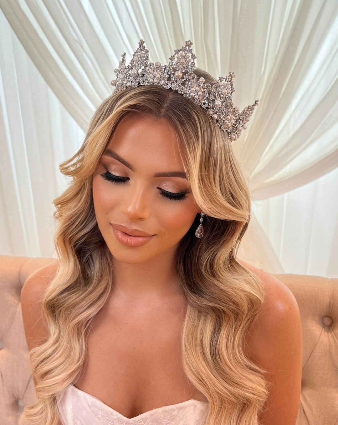 A woman with wavy blonde hair wears a sparkling Regal Bridal Crown style no. 80038 and teardrop earrings. She has soft makeup with long lashes and nude lips against draped white curtains. Her serene expression complements her tube-style gown, embodying elegance and grace.