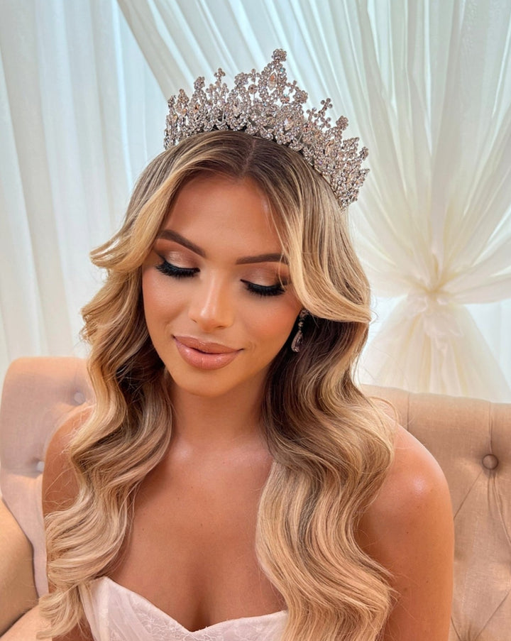 A woman with long, wavy blonde hair wears the exquisite Royal Wedding Tiara style no. 80034 and elegant makeup. Shes seated against a soft, cream-colored backdrop in a white top, looking downward with a gentle smile.