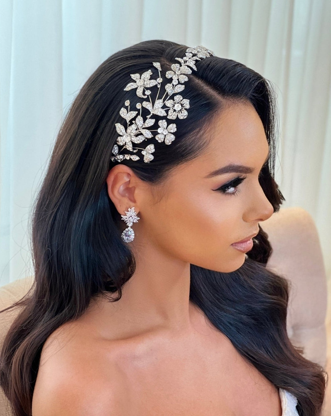 A woman with long, dark hair styled in loose waves wears the Asymmetrical Floral Swarovski Crystal Bridal Headband style no. 242687. Her makeup is natural, and shes seated against a light curtain backdrop.