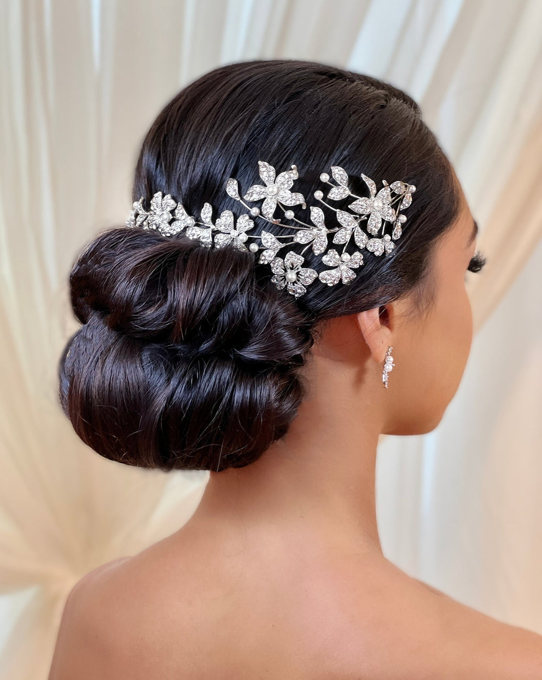 A woman with a sleek, dark updo hairstyle is adorned in the sparkling Asymmetrical Floral Bridal Headband (style no. 242687). Her earrings match its intricate design against a soft, light curtain backdrop.