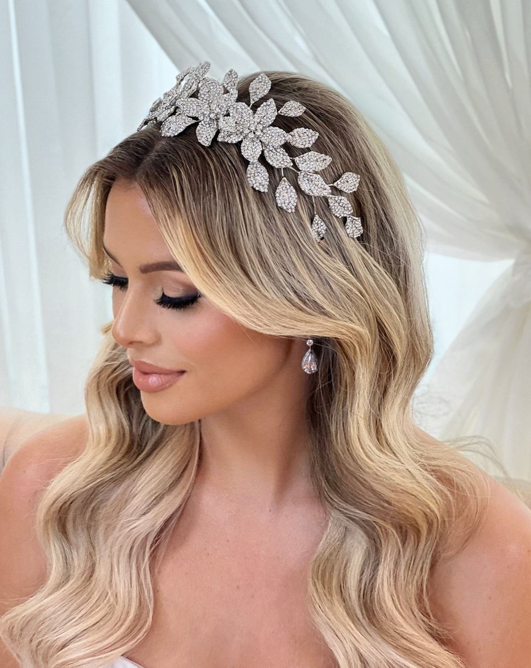 A woman with wavy blonde hair dons a stunning Crystal Flower Headband style no. 80070 and matching earrings. Her elegant makeup features smoky eyeshadow, long lashes, and nude lipstick, set against a softly draped white fabric background.