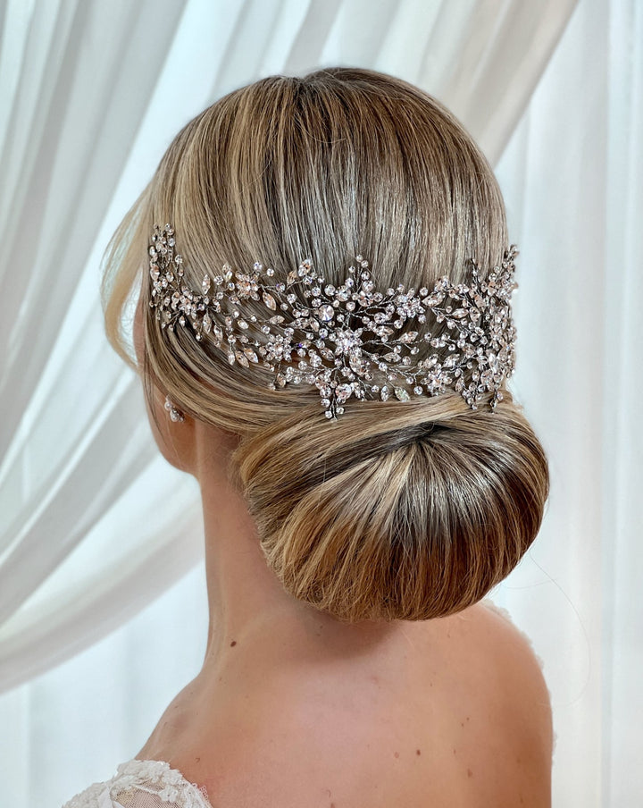 A woman with blonde hair styled in a low bun wears a Shimmering Swarovski Crystal Bridal Hair Vine, style no. 242480. It glistens with silver and crystal detailing. Sheer white curtains elegantly frame the background of the scene.