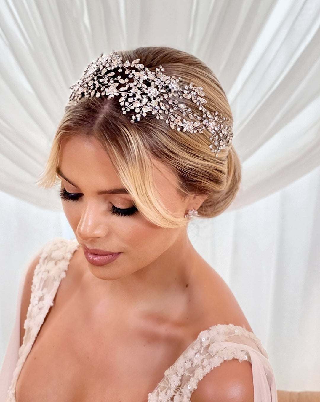 A woman with blonde hair styled in an elegant updo adorned by the Shimmering Bridal Hair Vine style no. 242480, set against a soft white draped background. She wears a sleeveless lace wedding dress and gazes down with her eyes gently closed.