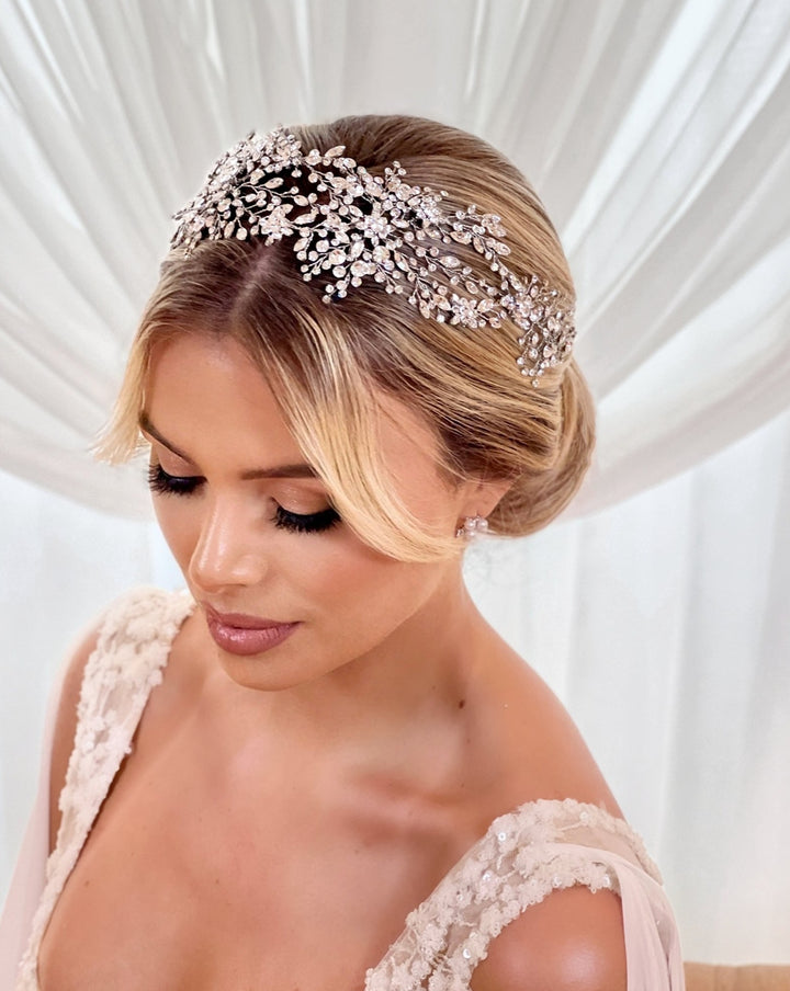 A woman with blonde hair styled in an elegant updo adorned by the Shimmering Bridal Hair Vine style no. 242480, set against a soft white draped background. She wears a sleeveless lace wedding dress and gazes down with her eyes gently closed.