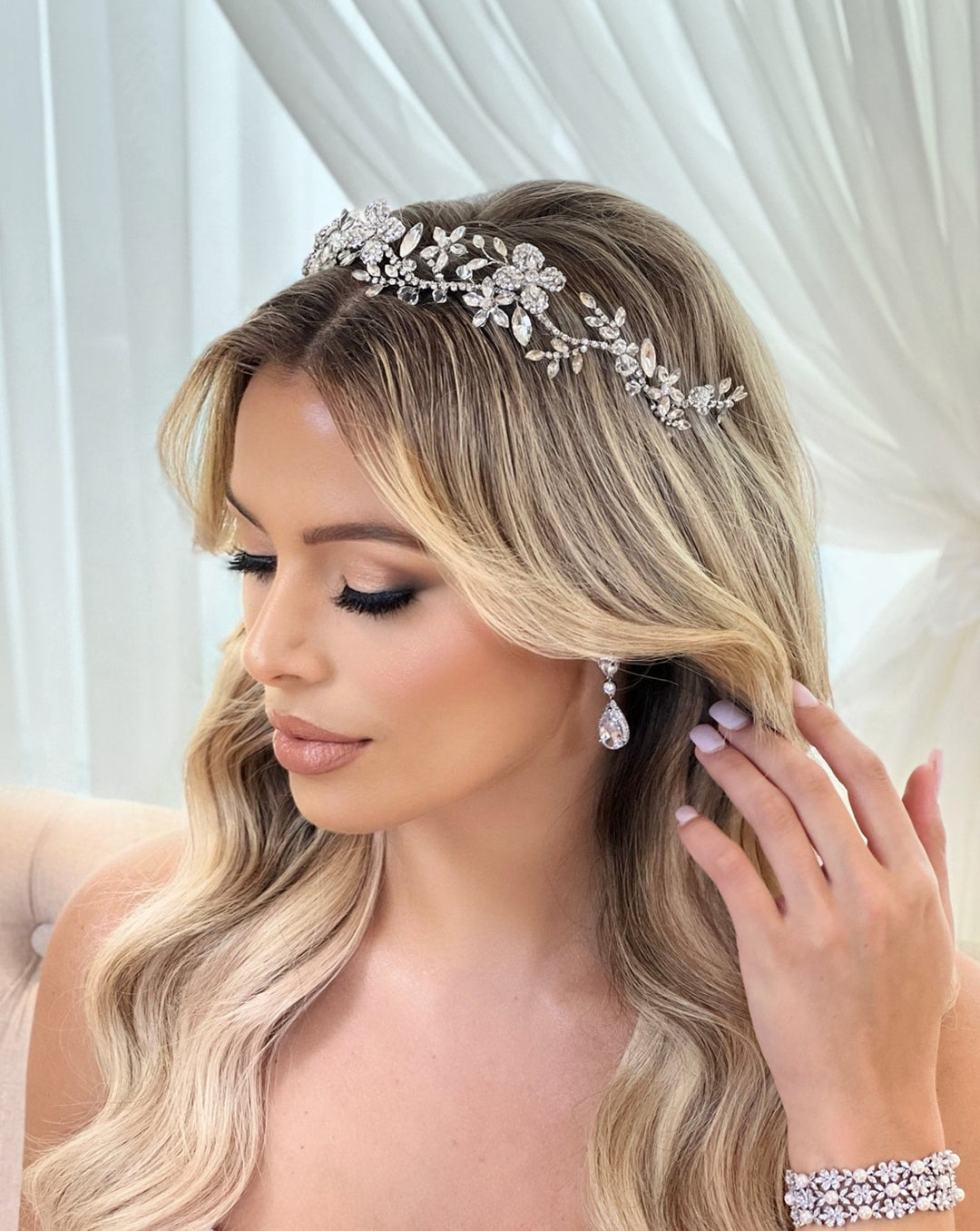 A woman with long blonde hair styled in loose waves wears a sparkling Delicate Floral Headband style no. 242694 and Swarovski hair vine alongside drop earrings. She has glamorous makeup and is adjusting her hair while looking down, seated before sheer white curtains.