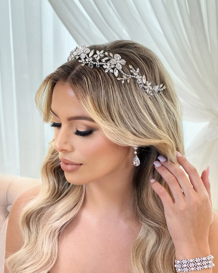 A woman with long blonde hair styled in loose waves wears a sparkling Delicate Floral Headband style no. 242694 and Swarovski hair vine alongside drop earrings. She has glamorous makeup and is adjusting her hair while looking down, seated before sheer white curtains.