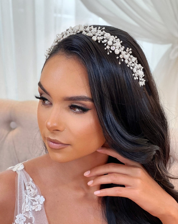 A woman with long, dark hair adorned with the Dramatic Asymmetrical Pearl and Swarovski Crystal Vine style no. 242703 gazes thoughtfully. She wears a delicate lace dress, seated on a tufted chair against a soft draped backdrop, creating an elegant and serene atmosphere.