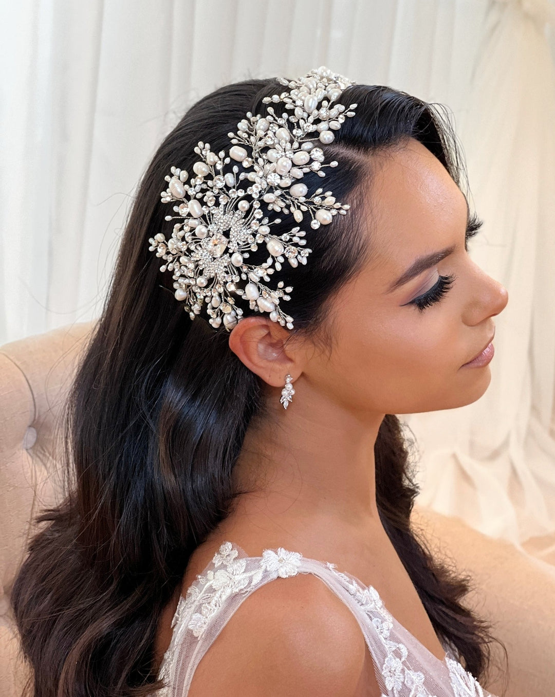 A woman with long dark hair, eyes closed, sits sideways wearing a delicate lace dress and the Dramatic Asymmetrical Pearl and Swarovski Crystal Bridal Hair Vine (style no. 242703). The background softly blurs with hints of white fabric.