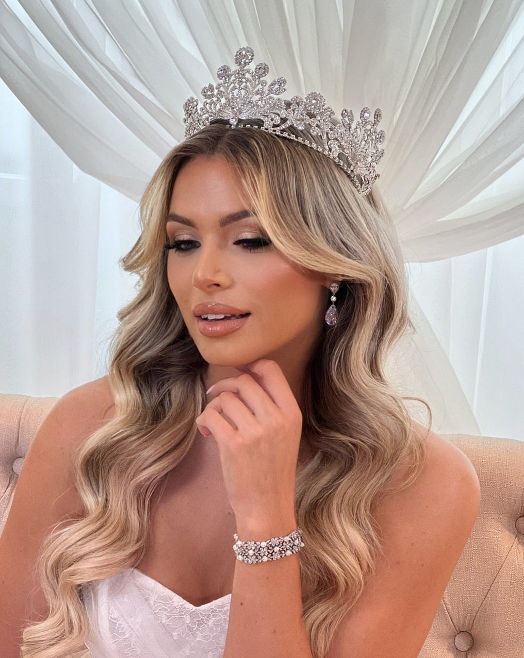 A person with long, wavy blonde hair wears a sparkling Classical Swarovski Bridal Crown style no. 80097 and matching jewelry, including earrings and a bracelet. Theyre in front of sheer white curtains, dressed in white, and gazing down thoughtfully.