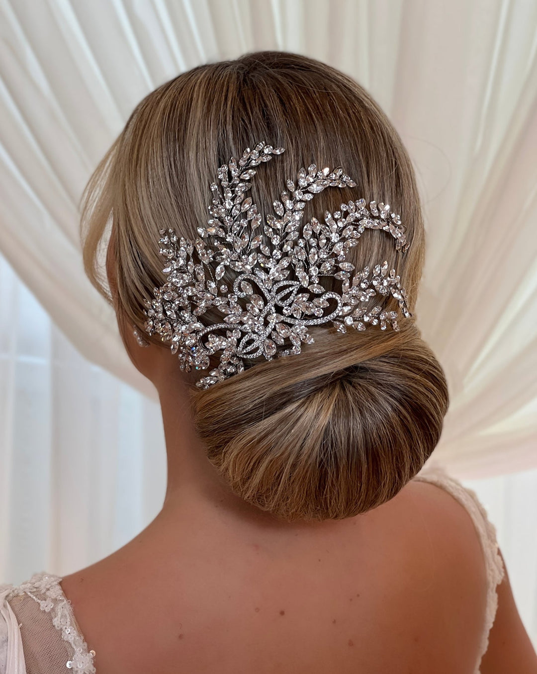 A woman with blonde hair styled in an elegant low bun is adorned with the Glamorous Crystal Bridal Comb (style no. 242535), featuring intricate Swarovski crystal designs. She pairs it gracefully with a delicate lace dress against a softly lit cream curtain backdrop.