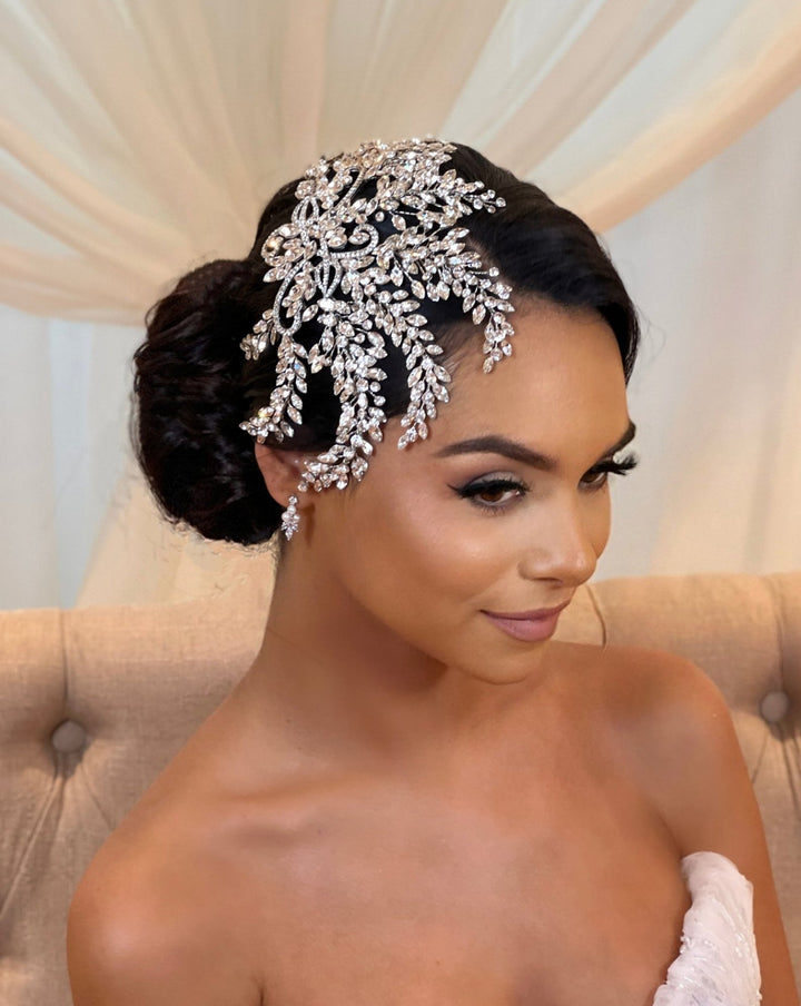 A woman with dark hair styled in an elegant updo adorned with the Glamorous Swarovski Crystal Hair Comb (style no. 242535). She wears subtle makeup and small earrings while seated on a beige chair against a softly draped curtain backdrop.