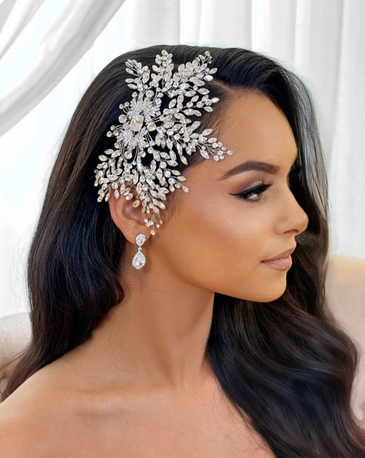 A woman with flowing dark hair wears a Dazzling Crystal Bridal Comb (style no. 242579) adorned with Swarovski crystals, resembling a grand wedding crown. She has teardrop earrings and poses against a light background, looking off to the side.