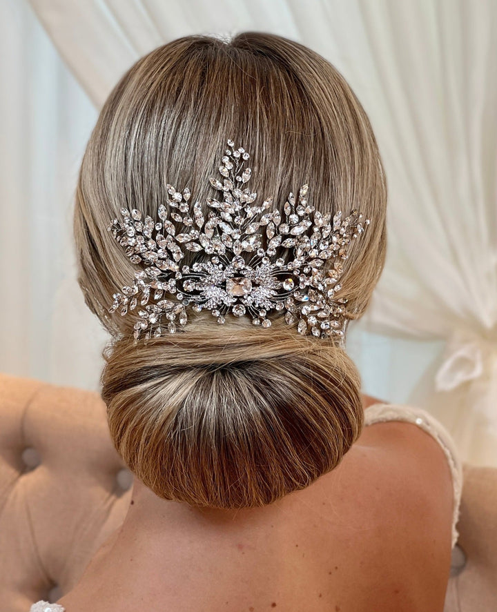 A woman's hair styled in an elegant low bun is accented with the Dazzling Swarovski Crystal Hair Comb, style no. 242579, featuring intricate floral and leaf patterns. Light-colored drapes and a tufted chair in the background enhance the serene atmosphere.