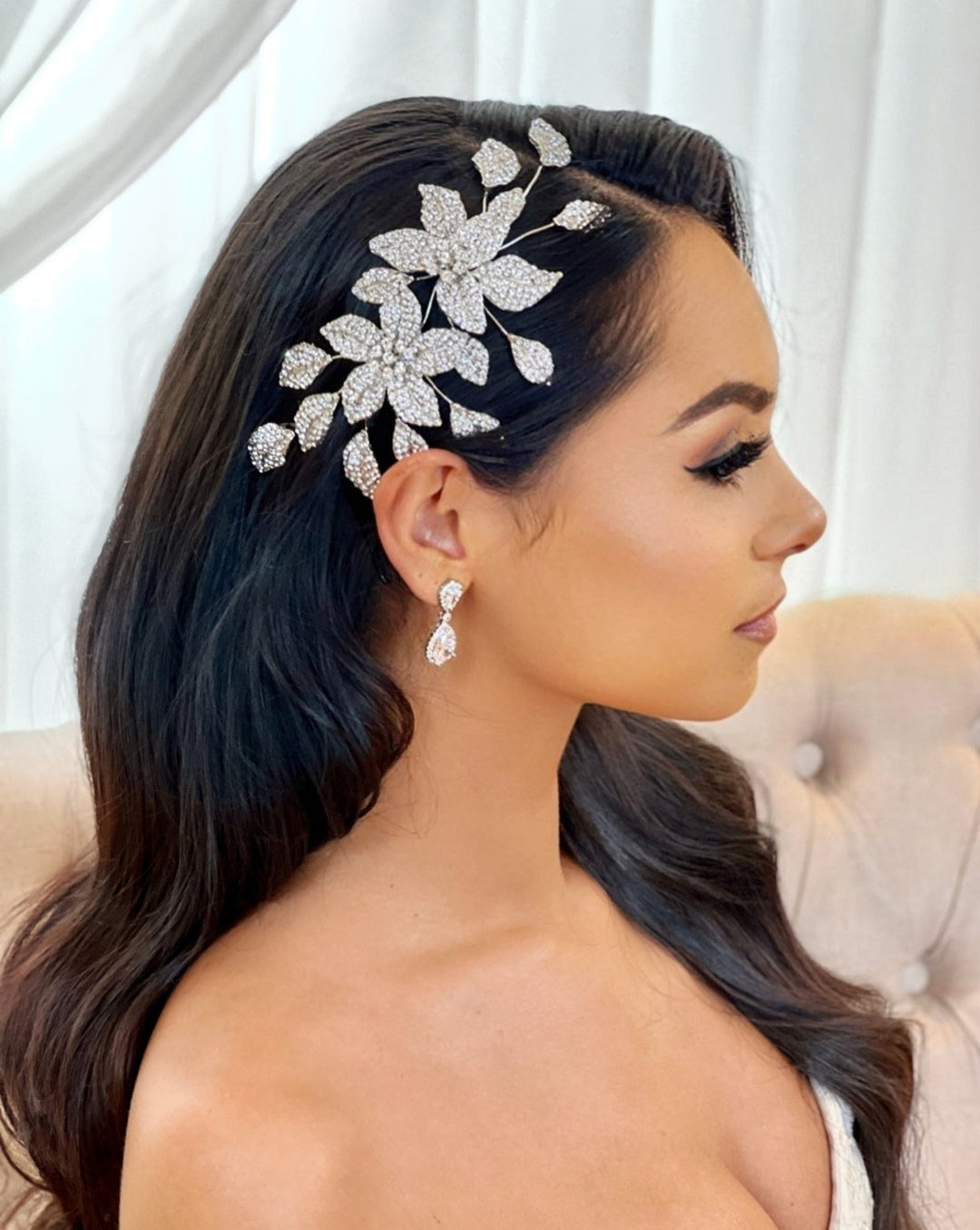 A woman with long, dark hair wears a Crystal Double Flower Comb (style no. 80091) and matching dangling earrings, seated on a tufted chair in profile against a light background.