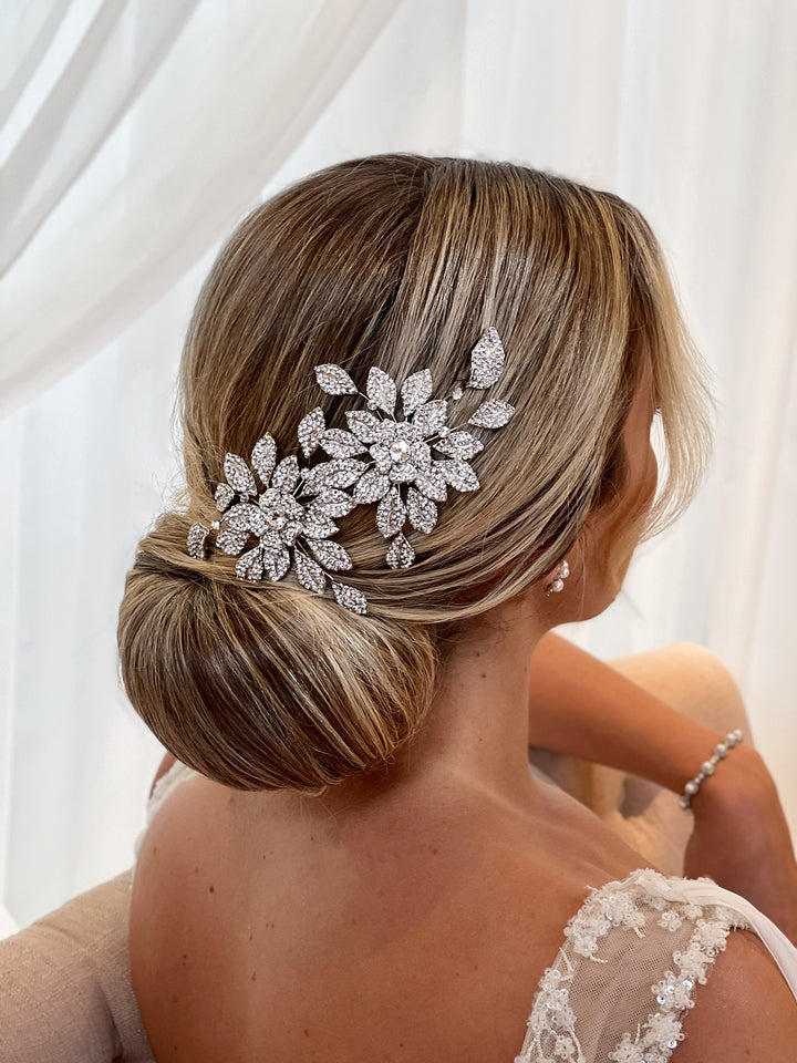A woman with blonde hair styled in a low bun, adorned with a Dazzling Double Blossom Comb style no. 80065  sits in front of a white curtain. Shes wearing a lace dress and bracelet, looking elegantly to the side.