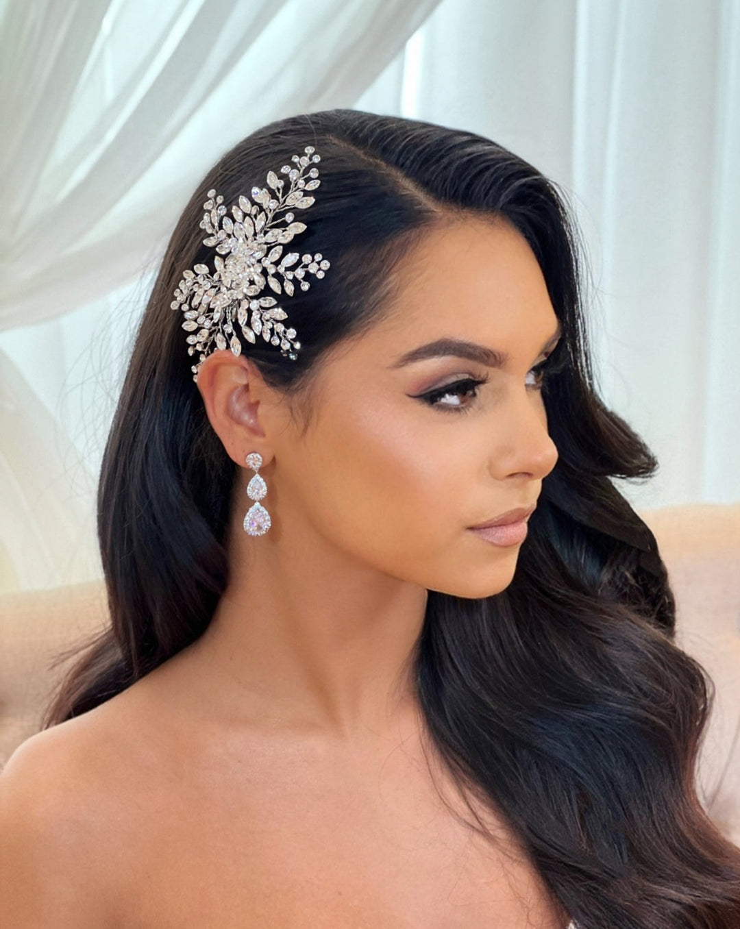 A woman with long dark hair wears an Elegant Bridal Hair Comb (style no. 242512) featuring stunning Swarovski crystals positioned on the side of her head. She has teardrop CZ bridal earrings and gazes softly to the side against an elegantly blurred backdrop of light-colored drapes.
