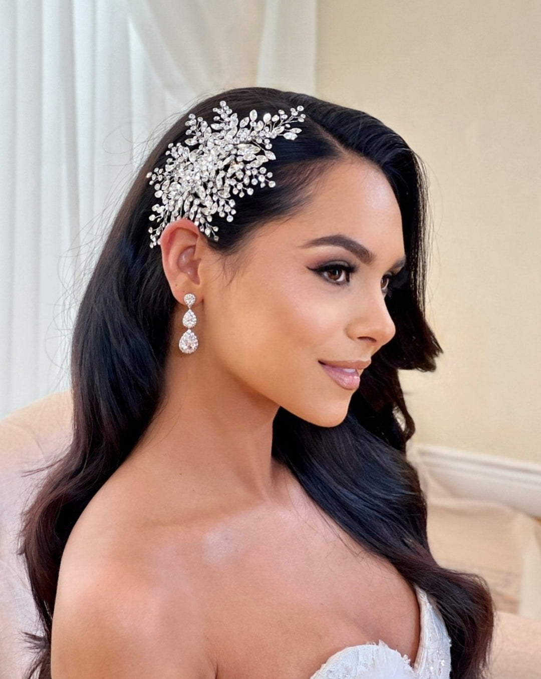 A woman with long dark hair wears an elegant and dramatic Swarovski bridal hair comb and matching earrings. Shes in a likely bridal white outfit against a neutral background, exuding serene elegance. On her head is the Soft Crystal Cluster Comb, style no. 242588.