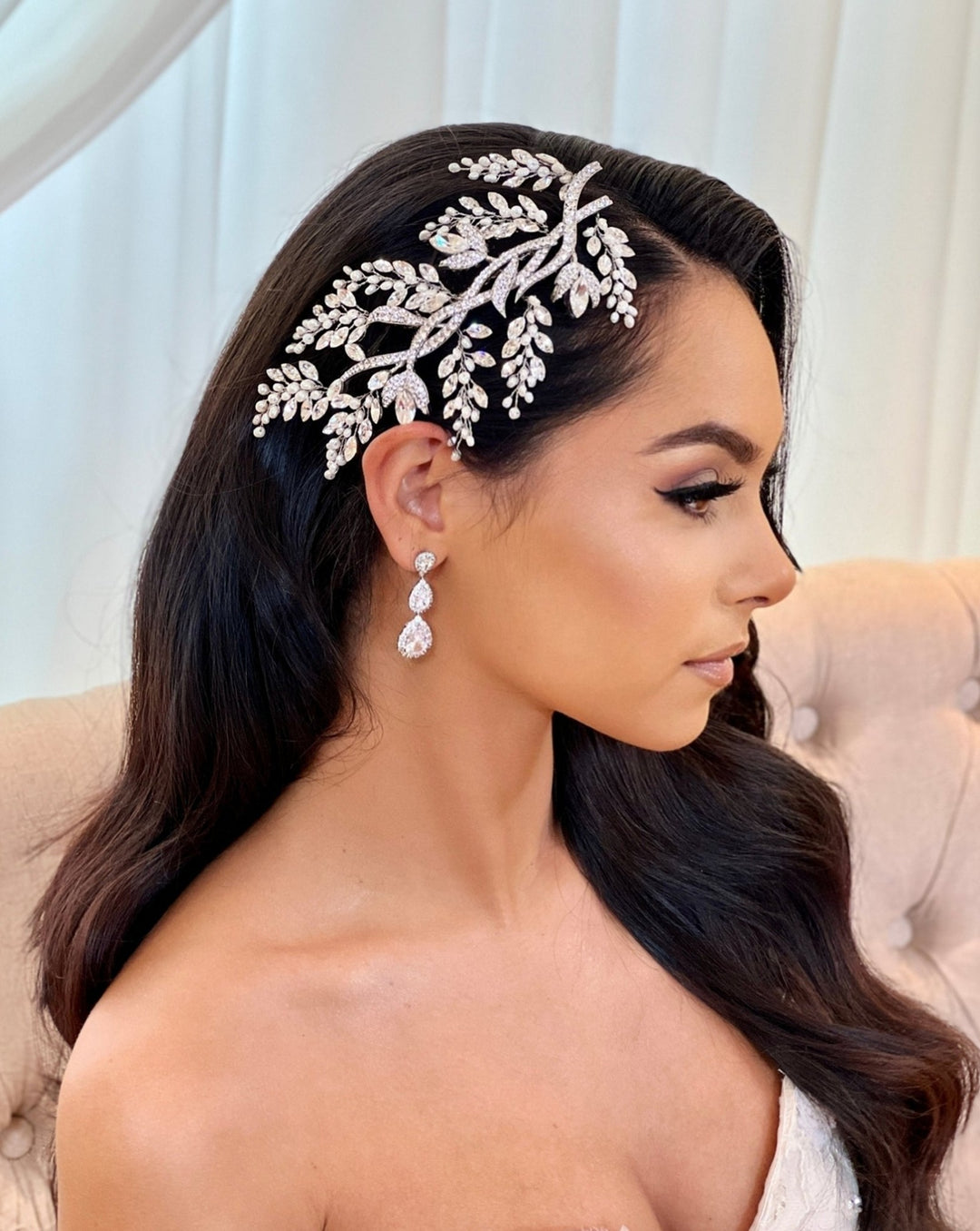 A woman with long dark hair is seated, wearing an Enchanted Forest Comb (style no. 242462) with a leaf design. She accessorizes her Swarovski bridal headpiece  with drop earrings and a strapless top. The softly blurred background of light-colored curtains and a cushion creates an elegant ambiance.