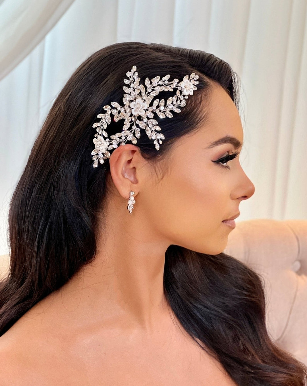 A woman with long, dark hair is shown in profile, wearing a Swarovski Crystal and Porcelain Flower Bridal Comb (style no. 242726), an ornate jeweled bridal hair accessory shaped like flowers and leaves. Her matching earrings are also visible against a softly blurred background, highlighting her elegant appearance.