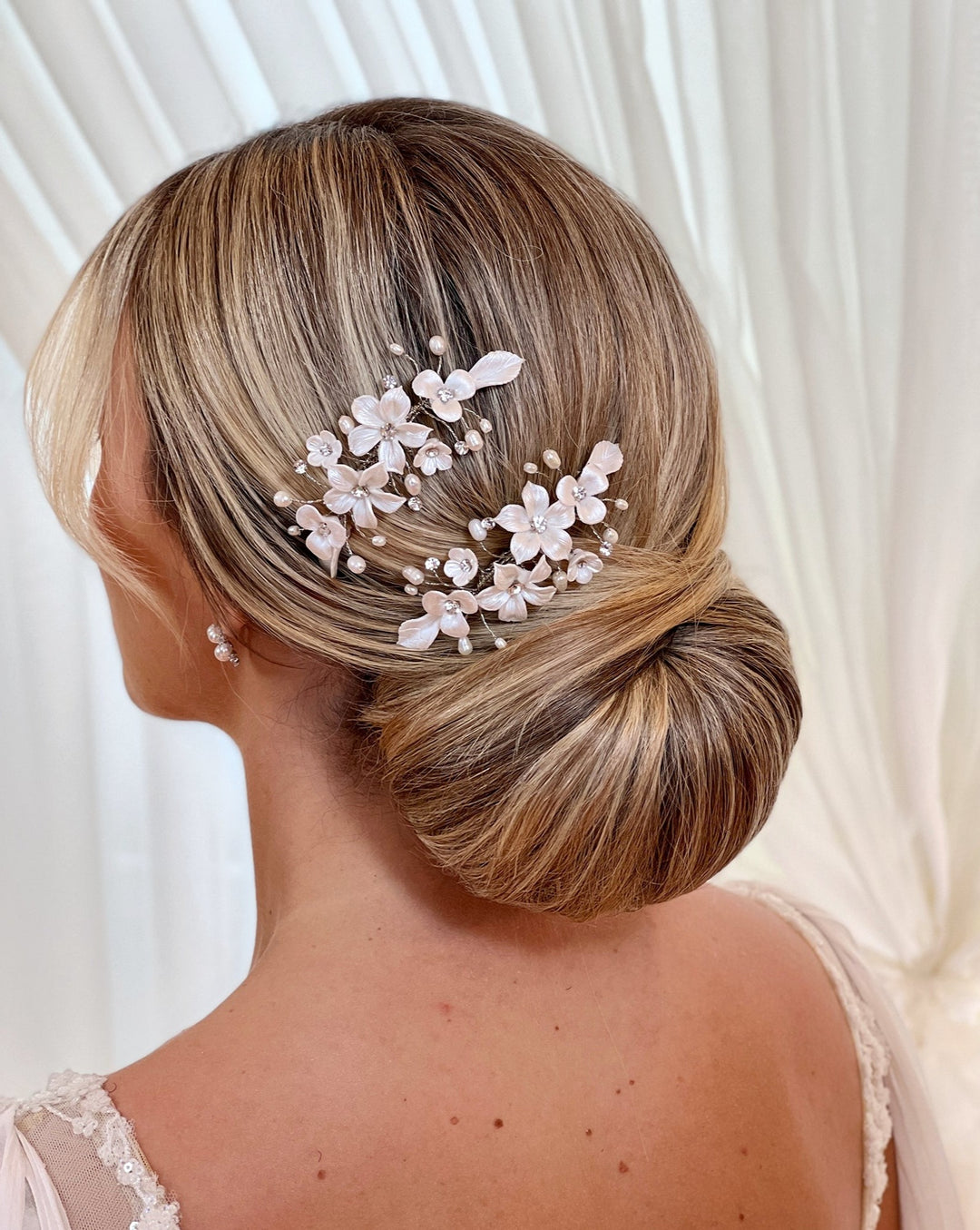 A woman with blonde hair styled in an elegant low bun adorned with the Romantic Porcelain Flower Hair Comb (style no. 242724). She is wearing pearl earrings against a soft, light-colored curtain background.