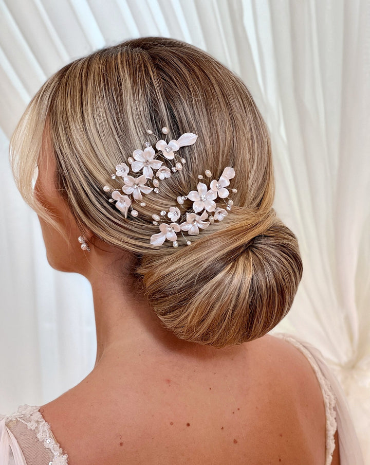 A woman with blonde hair styled in an elegant low bun adorned with the Romantic Porcelain Flower Hair Comb (style no. 242724). She is wearing pearl earrings against a soft, light-colored curtain background.