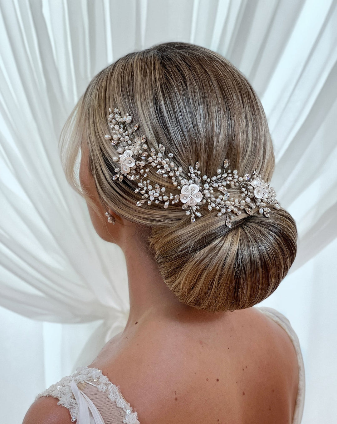 The image depicts a woman with a blonde low bun, adorned with the Romantic Pearl and Porcelain Flower Wedding Hair Vine (style no. 242714). She wears a white dress with sheer lace straps, set against a soft white backdrop.