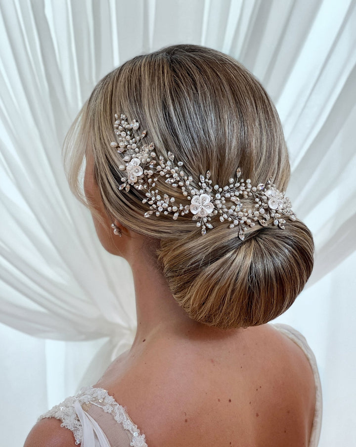 The image depicts a woman with a blonde low bun, adorned with the Romantic Pearl and Porcelain Flower Wedding Hair Vine (style no. 242714). She wears a white dress with sheer lace straps, set against a soft white backdrop.