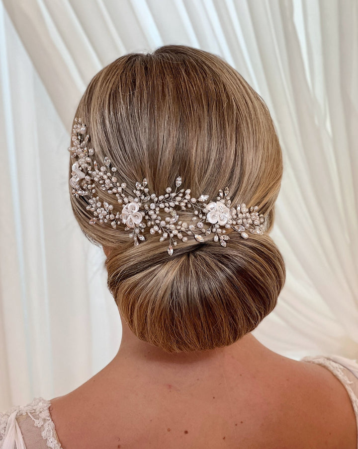 A woman with blonde hair styled in an elegant low bun adorned with the Romantic Pearl and Porcelain Flower Vine (style no. 242714) set against a soft white curtain backdrop.
