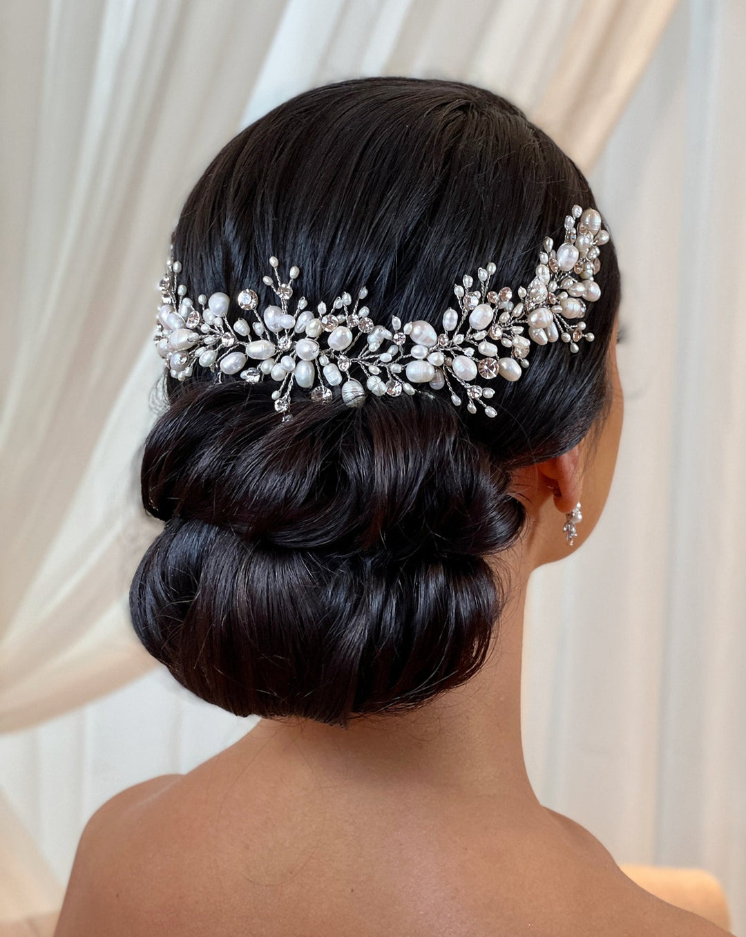 Close-up of a person with dark hair styled in a low bun, adorned with the Cluster Pearl and Swarovski Crystal Bridal Hair Vine (style no. 242704), set against soft draped fabric. They wear small, delicate earrings.