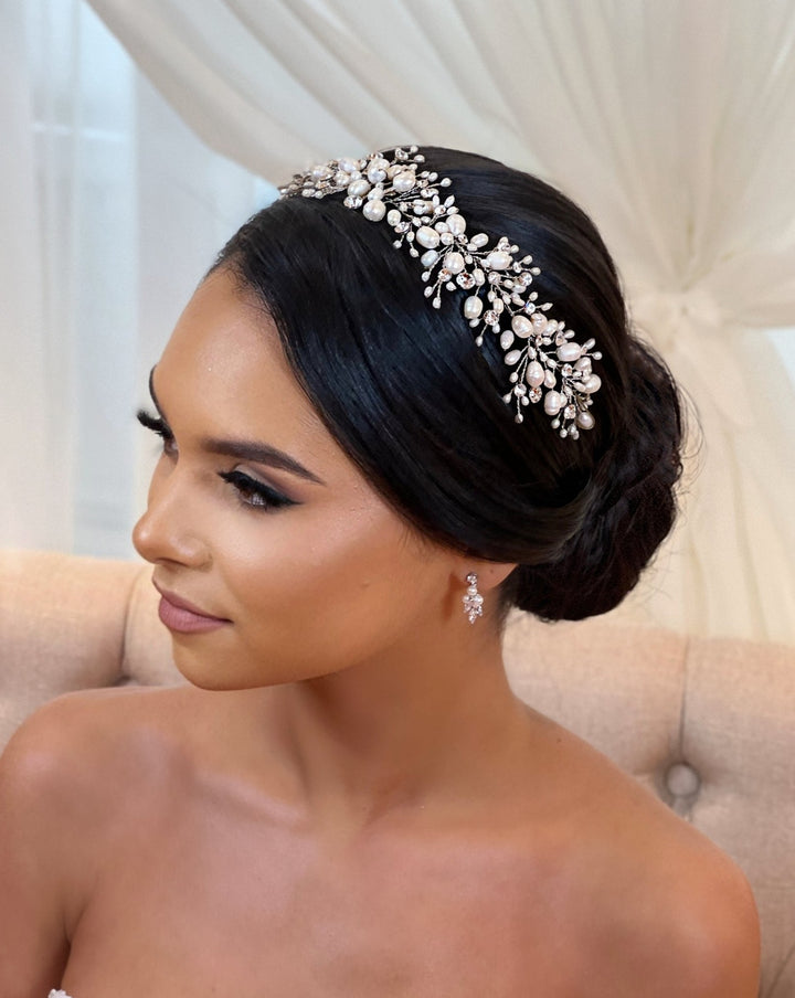 A woman with dark hair in an elegant bun wears a Cluster Pearl and Swarovski Crystal Bridal Vine (style no. 242704) with pearl earrings, seated against a light tufted background, exuding formal or bridal sophistication.