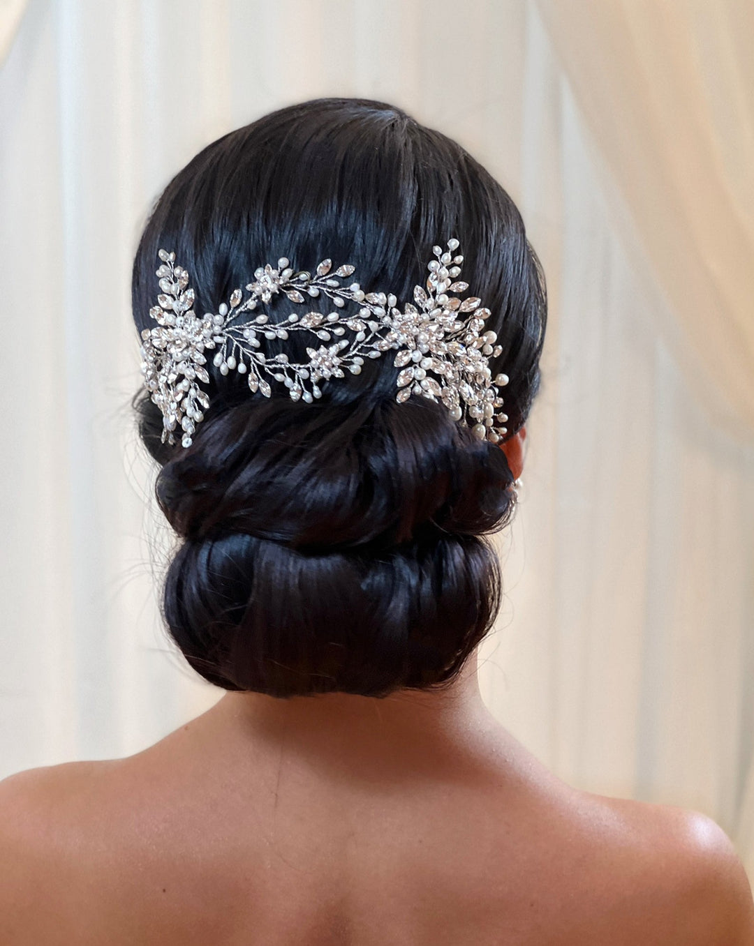 A woman’s hair is styled in an elegant, low bun adorned with the Stunning Crystal Bridal Hair Vine (style no. 242594), featuring intricate leaf and floral designs. The softly blurred background has light curtains.