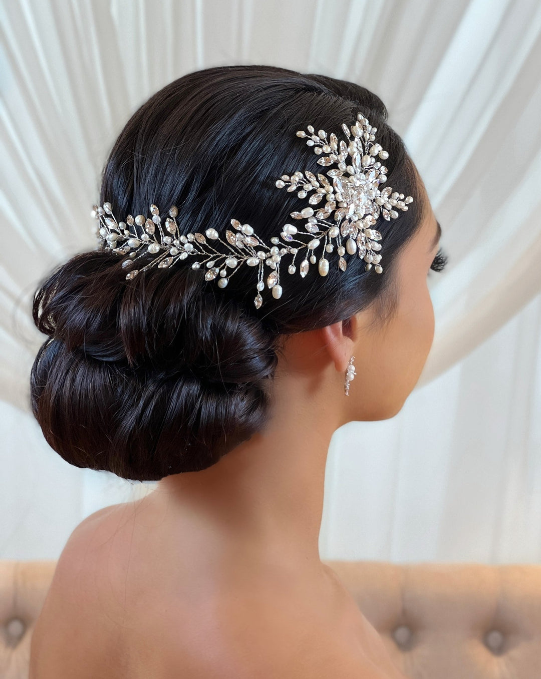 A person with dark hair styled in an elegant bun wears the Romantic Asymmetrical Vine style no. 242552. A Swarovski crystal bridal hair vine adorned with floral designs and pearls. Small matching earrings complete the sophisticated look.