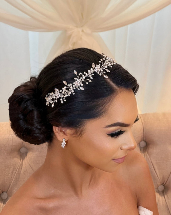 A woman with dark hair styled elegantly in a bun, adorned with the Sweet Pearl and Swarovski Crystal Hair Vine (style no. 242719), sits on a beige tufted sofa. She wears delicate earrings, complemented by her soft makeup. The sheer drapes form the subtle background.