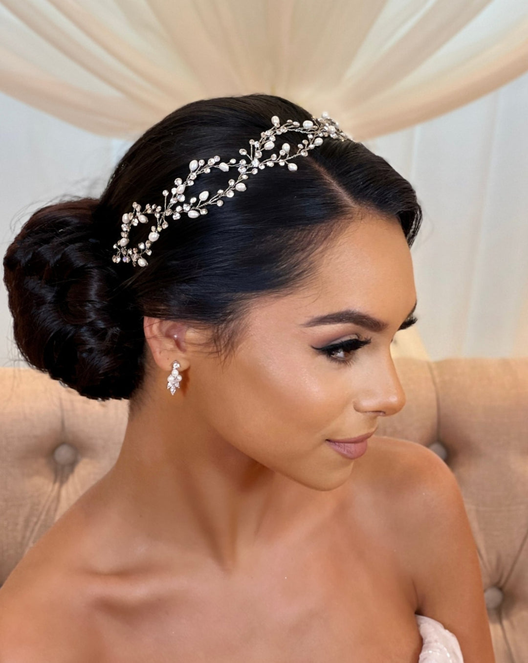 A woman with a low bun is adorned with the Elegant Looping Bridal Vine style no. 39228878. She complements it with drop CZ wedding earrings all set against a backdrop of soft, flowing white drapery.