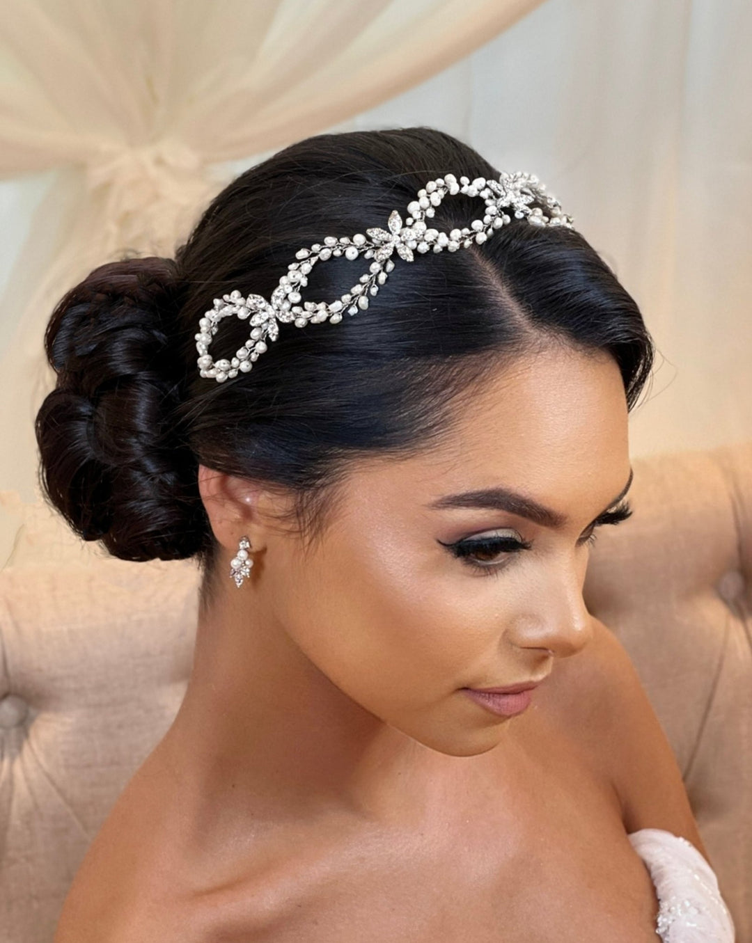 A woman with an elegant updo and pearl bridal earrings sits gracefully on a beige chair, her look enhanced by the Classy Pearl and Flower Looping Hair Vine style no. 242708. Her makeup is smooth, featuring winged eyeliner and soft lipstick, all set against a softly lit background.